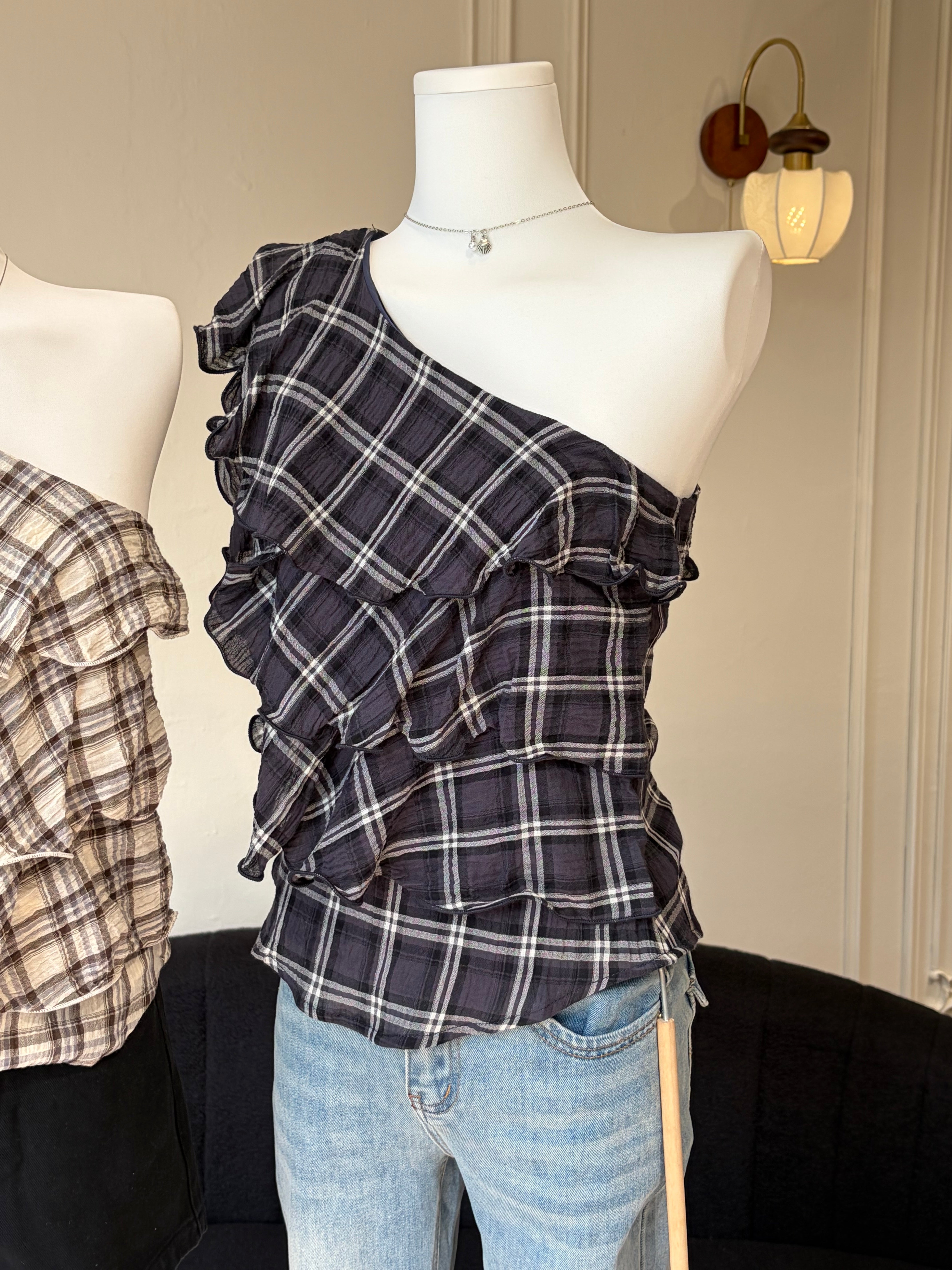 A dark navy and black plaid one-shoulder ruffled top displayed on a white mannequin torso, paired with light blue jeans. Another similar plaid top is partially visible on the left mannequin.