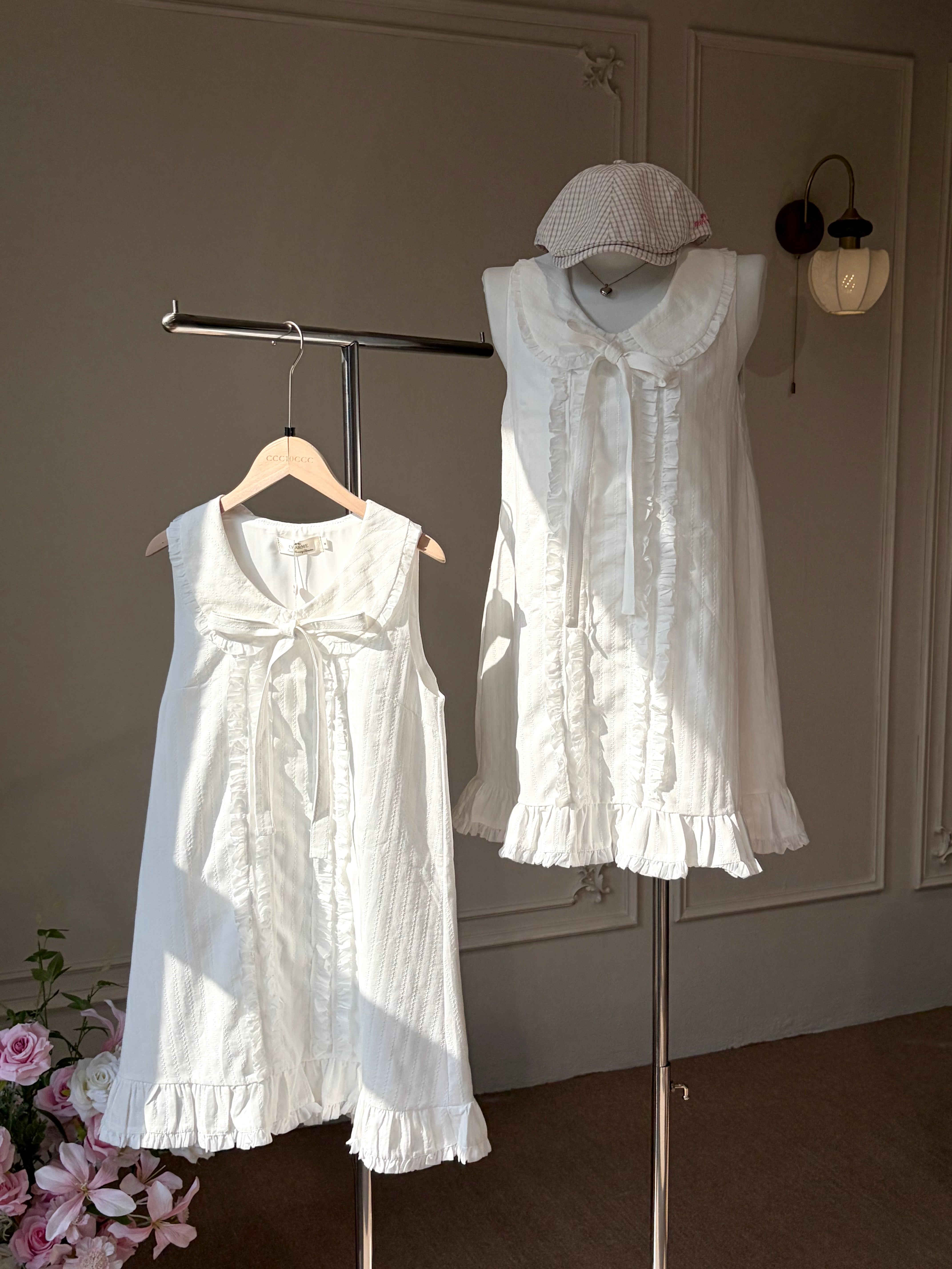Two white sleeveless baby dresses displayed on a clothing rack in an indoor room. One dress is shown on a wooden hanger, and the other is on a mannequin torso wearing a white bonnet.