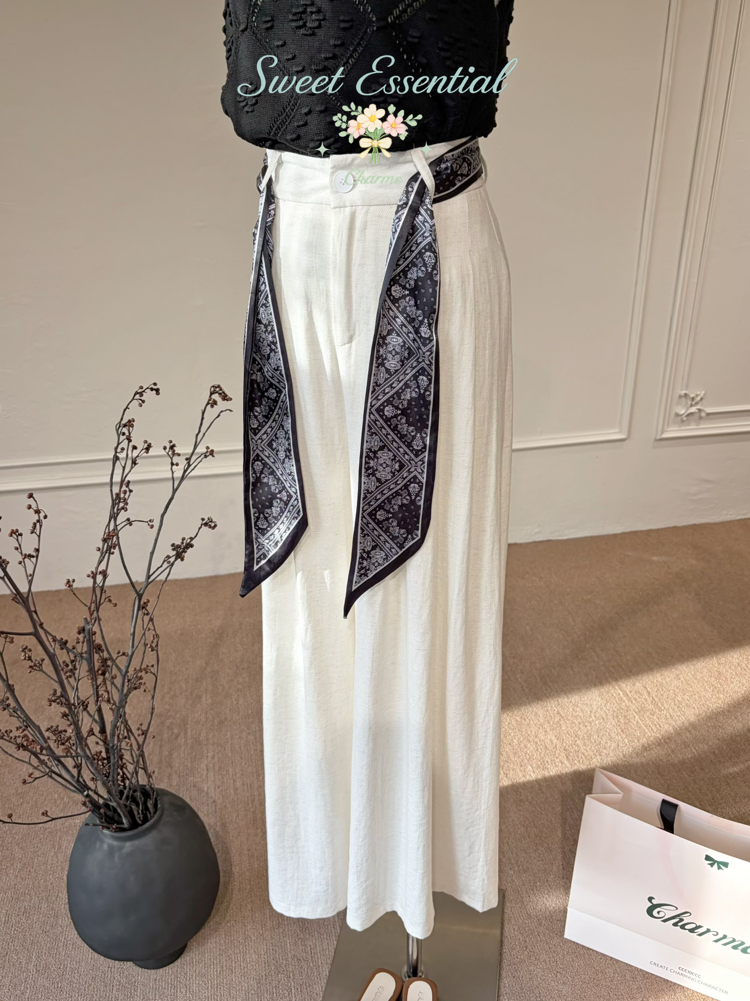 Person wearing a black top and white pants with a patterned scarf, standing in a room with a vase and a bag.