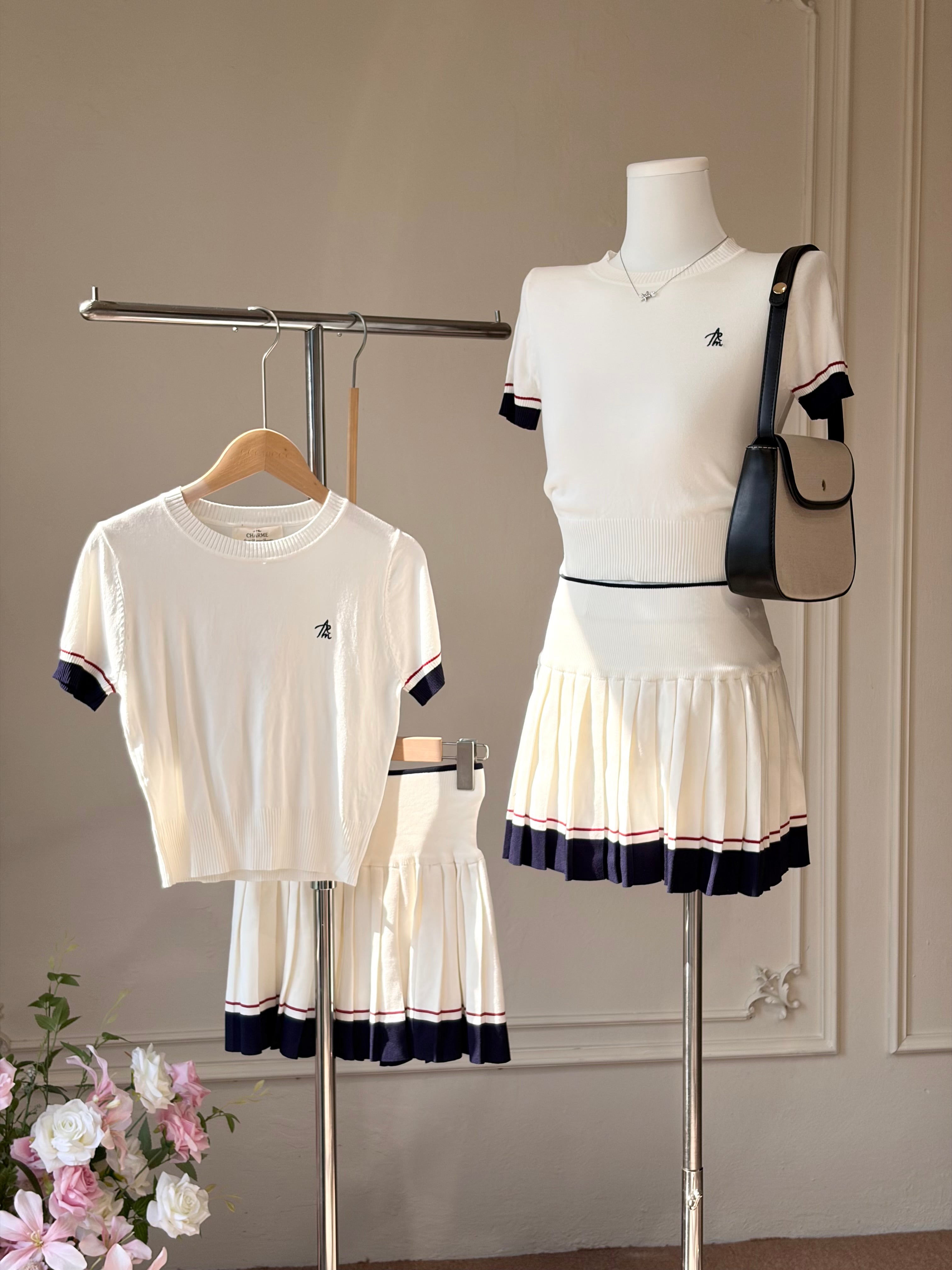 A coordinated women's outfit displayed on a mannequin and hanger against an indoor wall. The set includes a white short-sleeve cropped T-shirt with navy and thin red sleeve trim, a matching white pleated mini skirt with navy and red trim, and a small matching crossbody bag with a black strap and beige body.